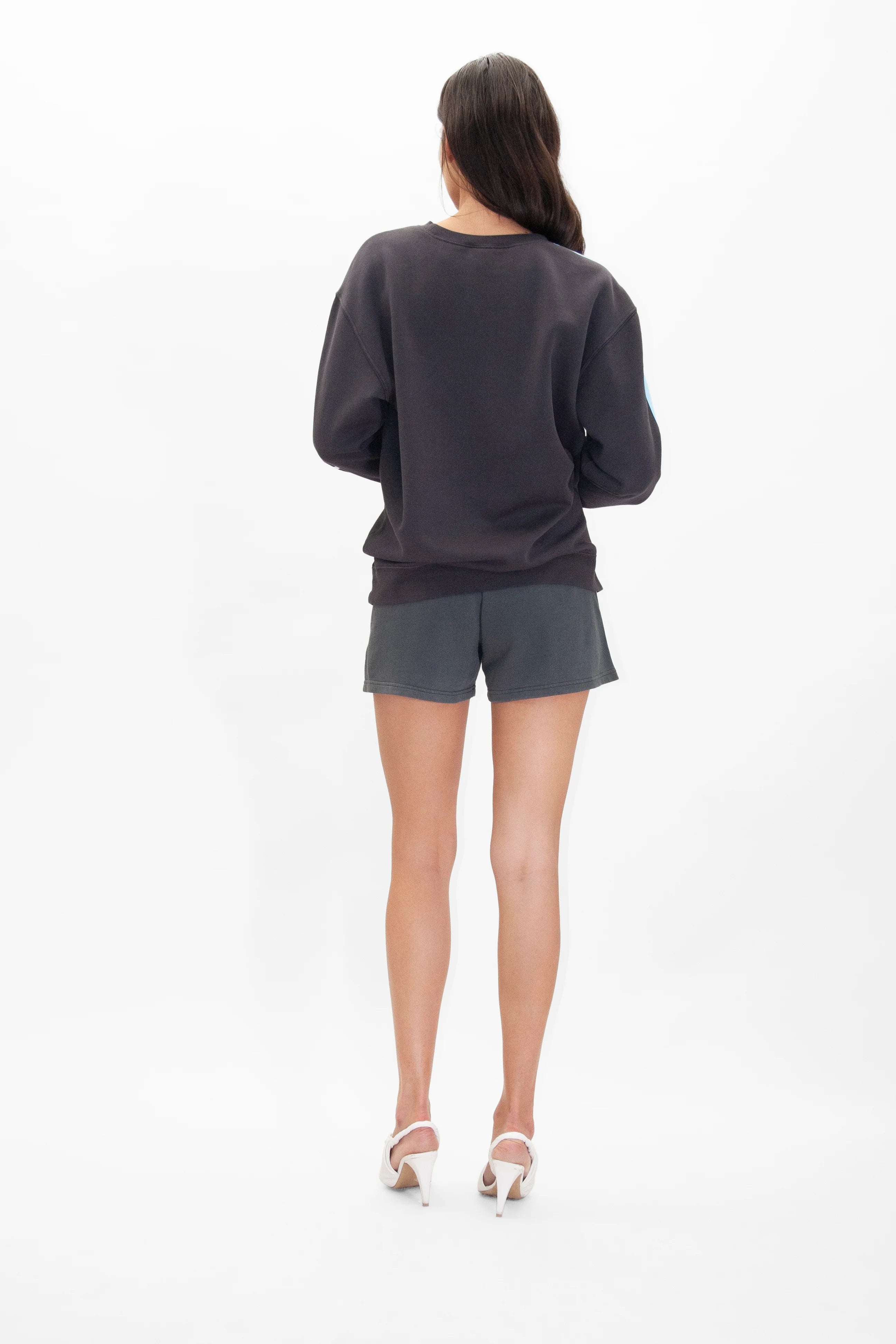 A person with long dark hair is standing with their back to the camera, wearing a GFLApparel Merkaba Crewneck in Space Glow, gray shorts, and white heels, against a white background.