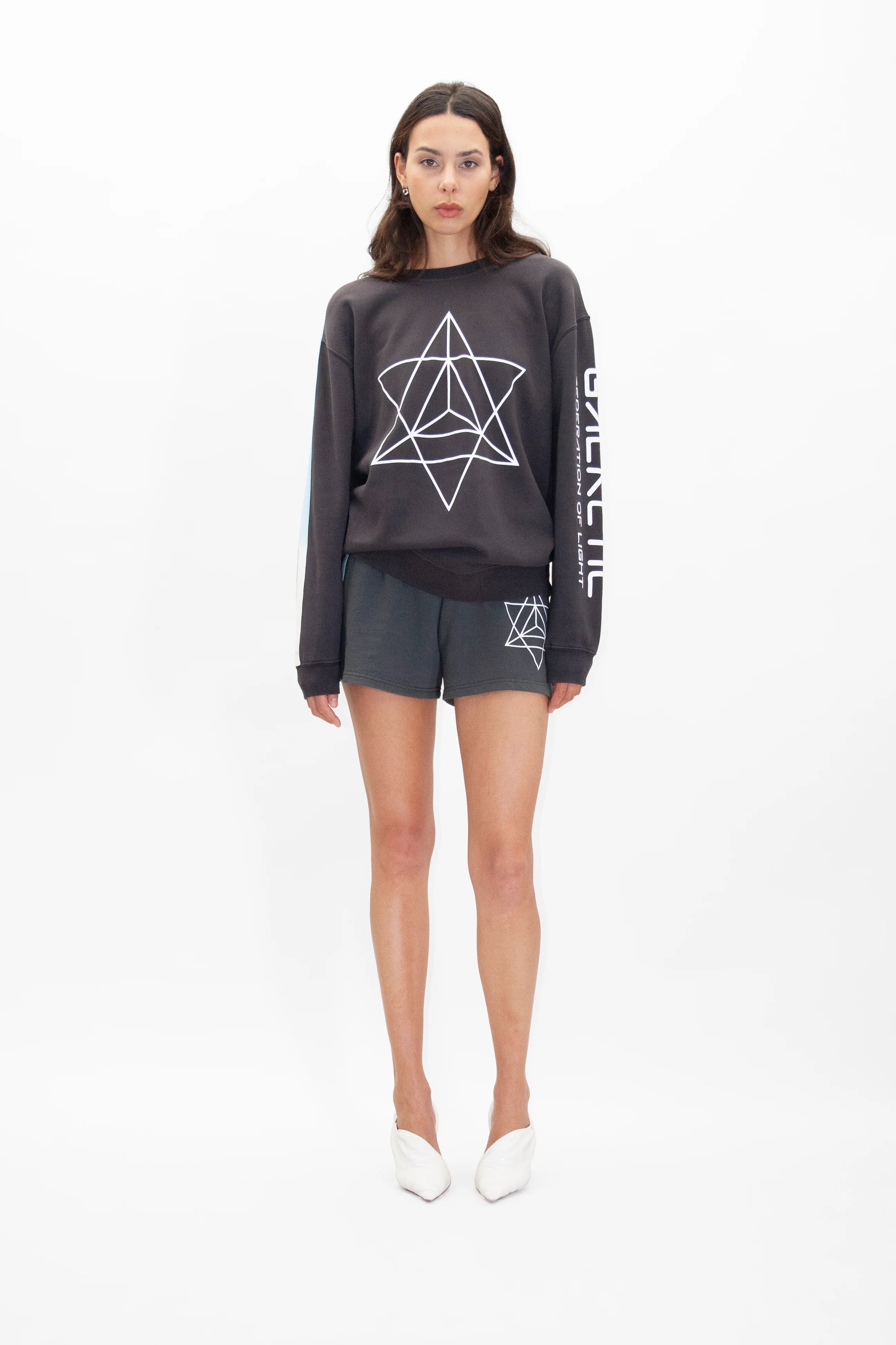 A woman stands against a white background, wearing a dark GFLApparel Merkaba Crewneck in Space Glow with a geometric star design and matching shorts. She has straight dark hair and is wearing white shoes.