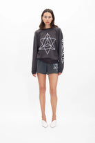 A woman stands against a white background, wearing a dark GFLApparel Merkaba Crewneck in Space Glow with a geometric star design and matching shorts. She has straight dark hair and is wearing white shoes.