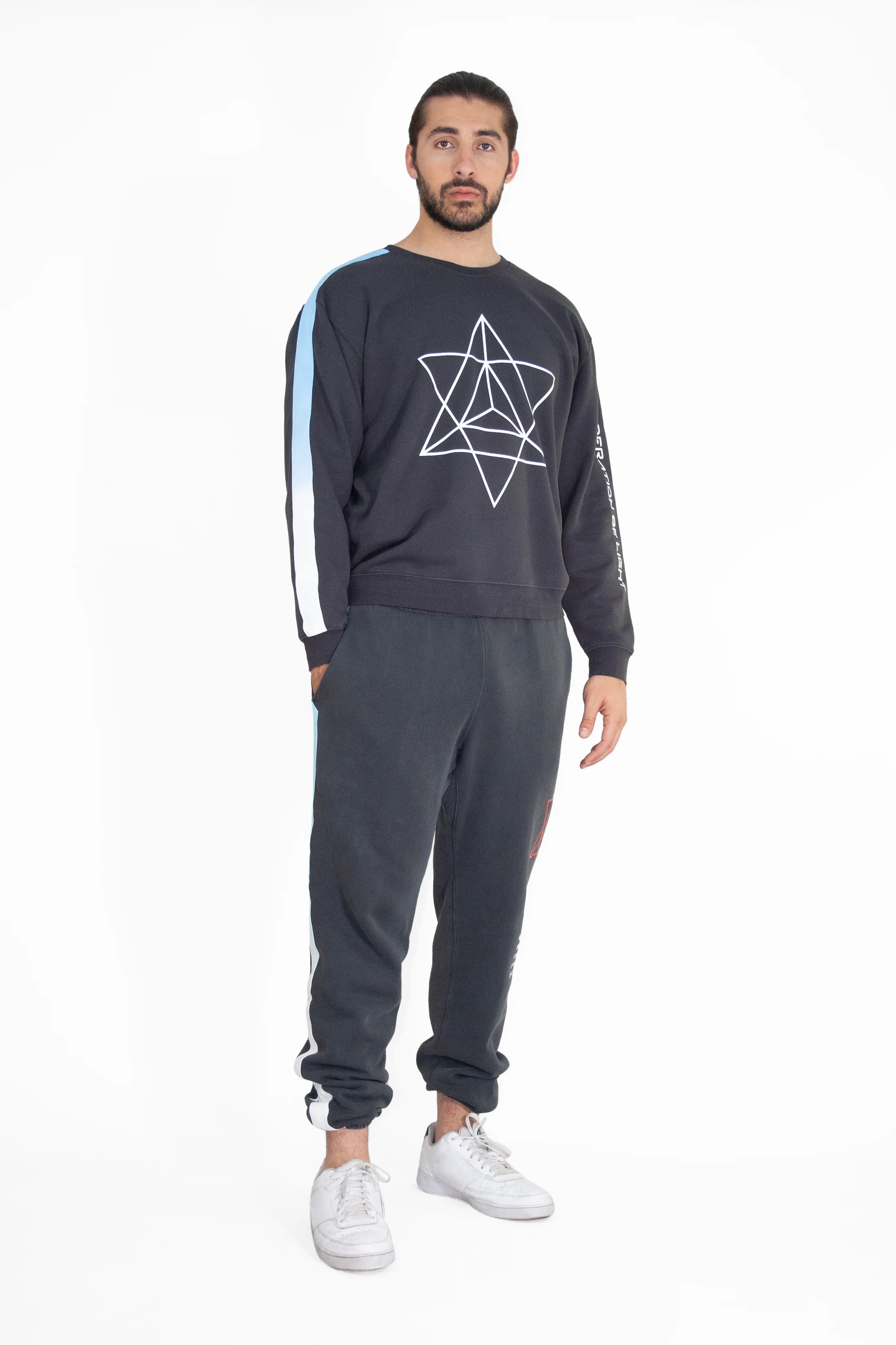 A man stands wearing a black GFLApparel Merkaba Crewneck in Space Glow featuring a geometric star design, dark sweatpants, and white sneakers. He has a neutral expression and is looking forward.