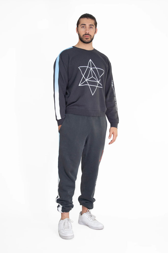 A man stands wearing a black GFLApparel Merkaba Crewneck in Space Glow featuring a geometric star design, dark sweatpants, and white sneakers. He has a neutral expression and is looking forward.