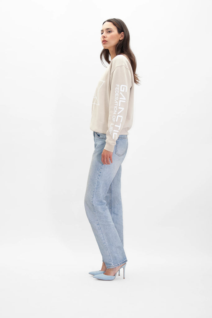 A woman in a beige long-sleeve shirt adorned with Sacred Geometry on the sleeve, light blue jeans, and blue high heels stands against a white background, facing slightly to the side. She is wearing the Merkaba Crewneck in Dune by GFLApparel.
