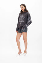 A woman stands against a white background, wearing a dark Merkaba Crewneck in Space Glow by GFLApparel with sacred geometry designs and the text "Galactic Federation" on the arm, paired with dark shorts and white heels.