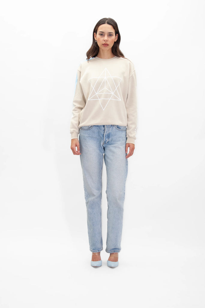A woman wearing a light-colored GFLApparel Merkaba Crewneck in Dune with a geometric design and light blue jeans stands against a plain white background.