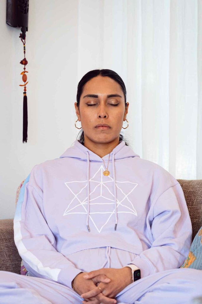 A person with closed eyes sits on a couch, wearing the GFLApparel Merkaba Cropped Hoodie in Nebula adorned with sacred geometry, including a Merkaba star design. With earrings and a smartwatch, the individual appears to be meditating.