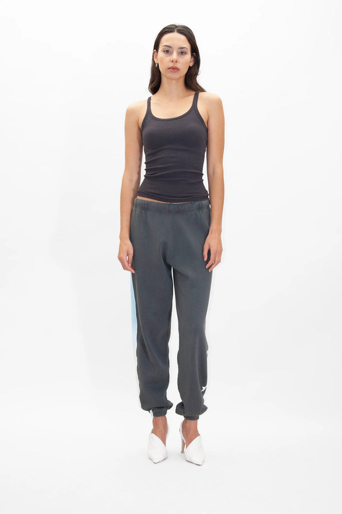 A woman stands against a plain white background, wearing a dark sleeveless top, GFLApparel GFL Stars Sweatpants in Space Glow with blue stripes, and white heels. She has long hair and maintains a neutral expression.