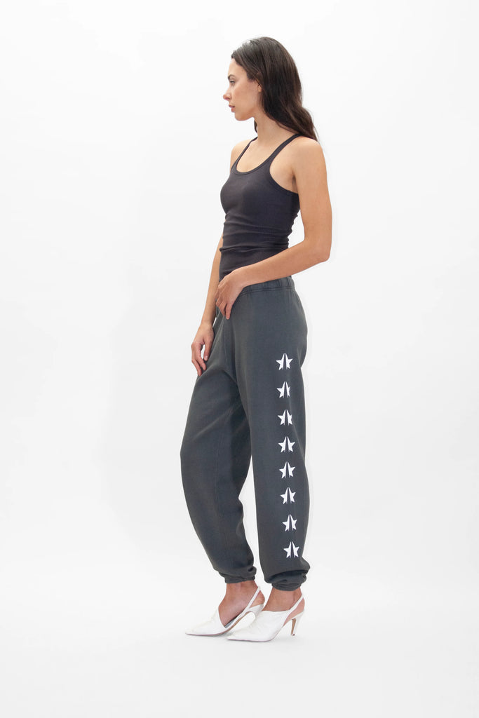 A woman in a black tank top and gray GFLApparel GFL Stars Sweatpants in Space Glow with white star designs on the side, standing in profile against a plain white background, showcasing her performance tech-ready outfit with white heeled shoes.