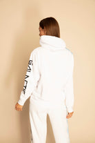 A person with long hair is turned away, wearing a white hoodie with "GFLApparel" printed on the sleeve, and matching white pants featuring subtle Sacred Geometry designs.