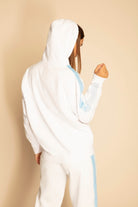 Person wearing a white and light blue GFLApparel Merkaba Hoodie in Lite Beam, facing away from the camera with right hand raised slightly, subtly revealing a pattern inspired by Sacred Geometry on the back of the hoodie, against a beige background.