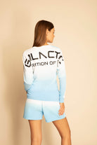 A person with long hair is standing, facing away, wearing a blue and white gradient crewneck-style ombré long-sleeve shirt and matching shorts. The breathable cotton shirt has large black lettering that is partially visible. They are wearing the GFL Stars L/S in Atmosphere from GFLApparel.