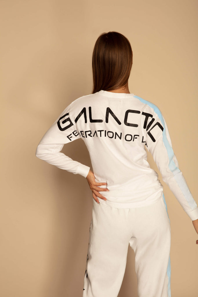 A person with long hair is standing with their back to the camera, wearing a white, long sleeve tracksuit adorned with "GALACTIC FEDERATION OF LOVE" and GFLApparel's GFL Stars L/S in Lite Beam printed on the back.