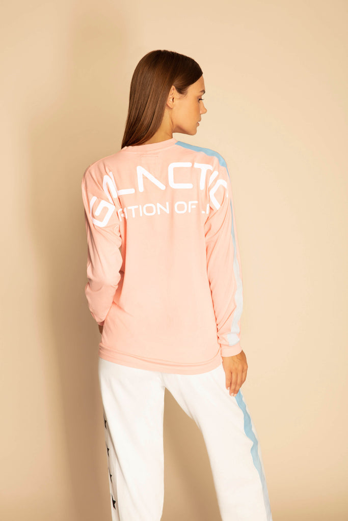 A woman with long brown hair wearing a pink GFLApparel GFL Stars L/S in Sun Fade made of breathable cotton and relaxed fit white pants with blue stripes is standing against a beige background.