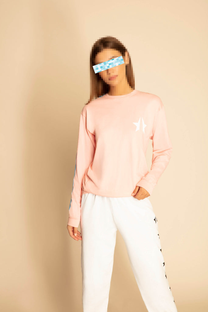 A person stands against a beige background, wearing a peach 369 L/S in Sunfade from GFLApparel with a white star emblem on the chest and white pants with black accents. The person's eyes are obscured with a pixelated effect, evoking an air of mystery akin to Nikola Tesla's enigma.