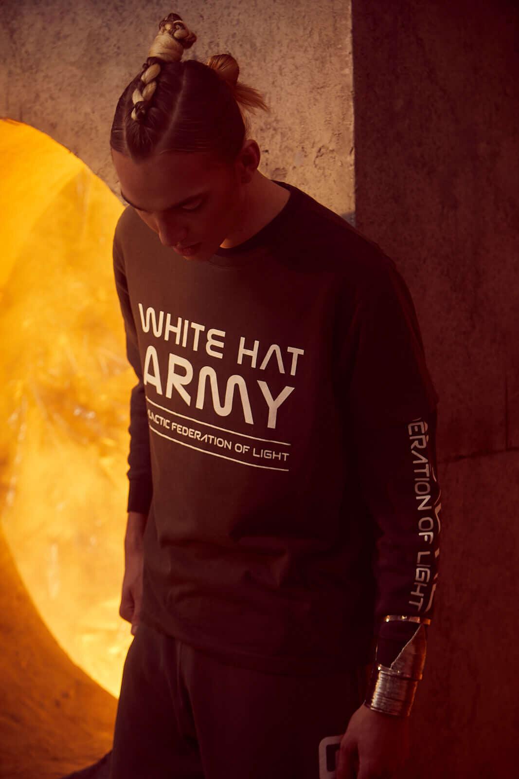 A person with braided hair wears a GFLApparel White Hat Army Tee in Caladan featuring a bold statement print: "White Hat Army - Galactic Federation of Light," and stands in front of an illuminated background.