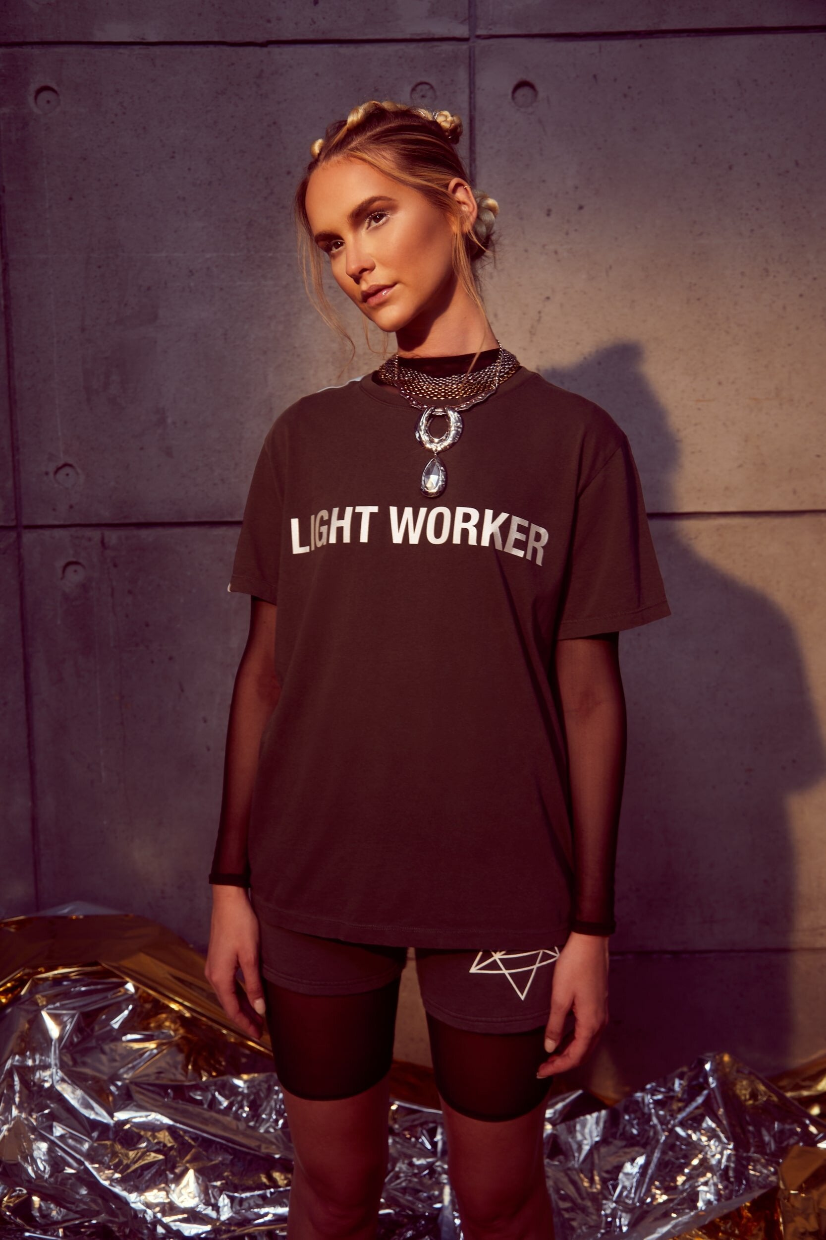 A person stands against a concrete wall wearing a dark GFLApparel Light Worker Tee in Space Glow with layered necklaces and shorts. The shirt’s reflective print adds a unique touch, and the breathable cotton ensures comfort. They appear in a casual yet stylish pose with metallic foil on the floor.