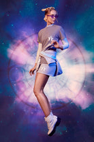A woman in futuristic attire stands against a vibrant, dreamy background of outer space, featuring a mix of blues, purples, and pinks with celestial elements. She's wearing bold print Star Shorts in Nebula from GFLApparel that complement the cosmic scene perfectly.