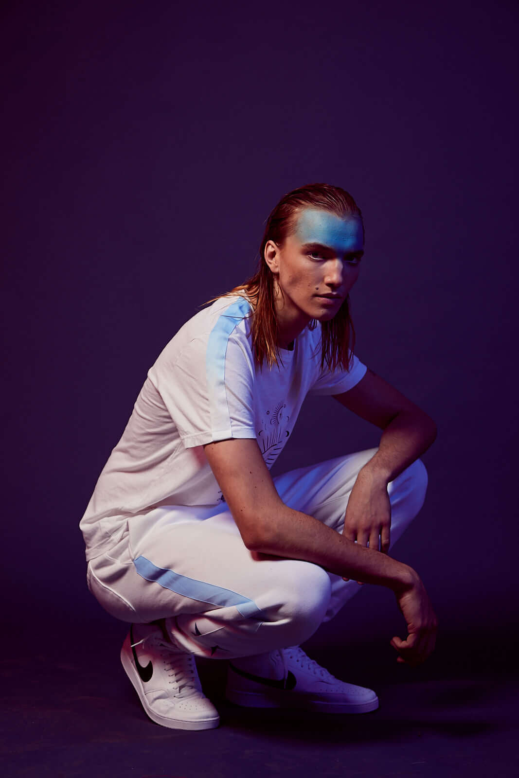 Person with long hair kneels, wearing a white outfit with blue accents and white sneakers, in a dimly lit setting with blue and purple lighting, the background faintly revealing patterns of Sacred Geometry, clad in the Moon Metatron Tee in Lite Beam by GFLApparel.
