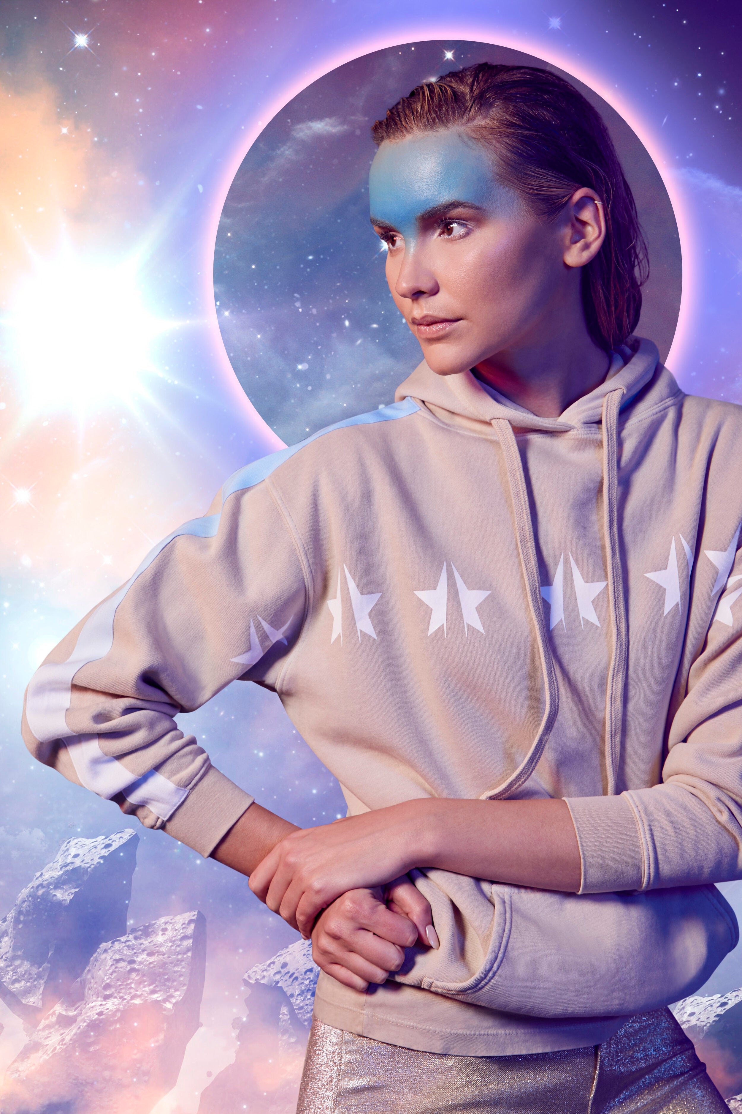 Person wearing a GFLApparel GFL Stars Hoodie in Dune with star designs and blue face paint, standing against a cosmic background with a bright burst of light and a halo-like circle.