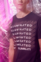 A person wearing an Illuminated Tee in Space Glow by GFLApparel, featuring the word "ILLUMINATED" in bold print repeated multiple times. The black shirt is made of breathable cotton. The background has a pink hue and appears slightly blurred.