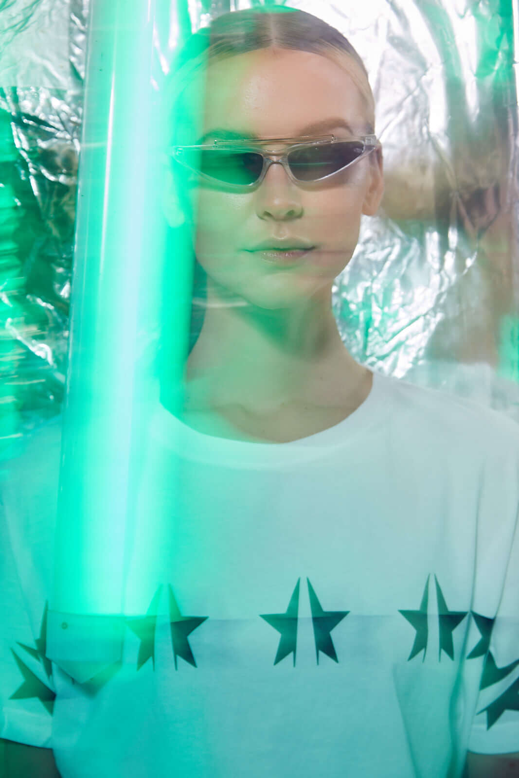 A person wearing sunglasses and a white GFLApparel GFL Stars Tee in Lite Beam made of breathable cotton with star patterns stands against a metallic background illuminated by green light.