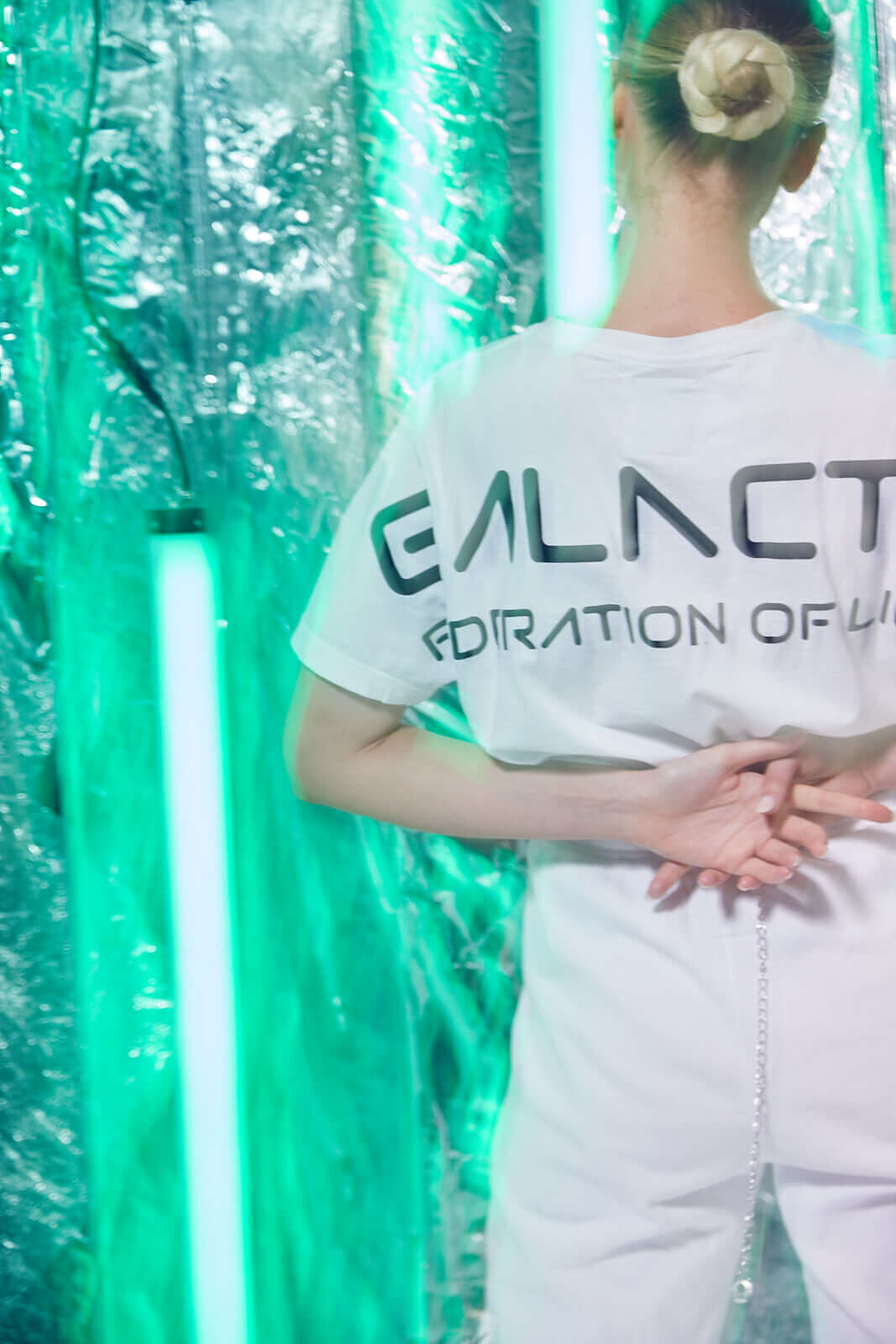 Person in a white GFLApparel GFL Stars Tee in Lite Beam with "GALACT F" written on the back, standing with hands behind their back in front of a green-lit futuristic backdrop. The relaxed fit and breathable cotton make it both stylish and comfortable.