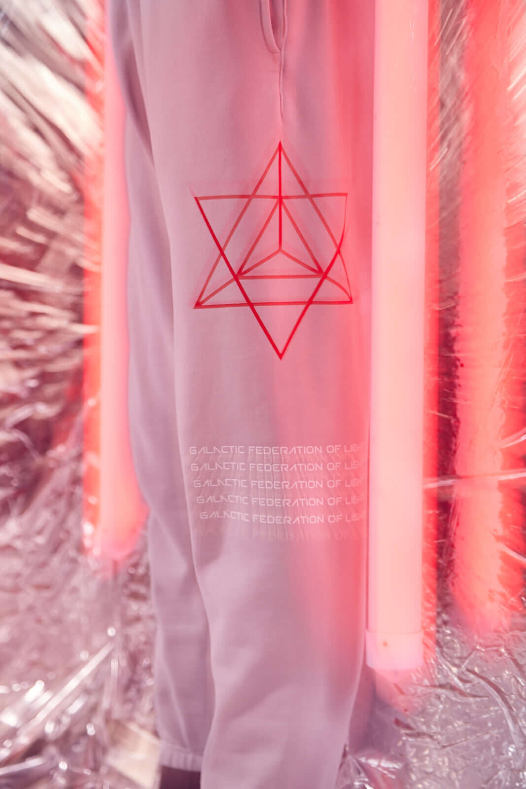 Person wearing GFLApparel Hypergalactic Pants in Galactic Gray with a geometric red design and "Galactic Federation of Light" text, featuring a drawstring waistband, standing near pink illuminated tubes against a metallic backdrop.