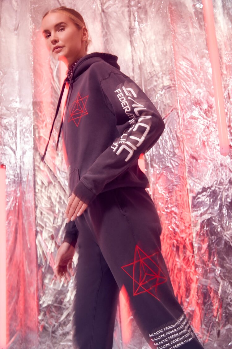 Person in a Merkaba Hoodie in Space Glow by GFLApparel, and matching pants with red sacred geometry designs, resembling a Merkaba, stands against a metallic backdrop with pink lights.