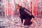 A person in a GFLApparel Merkaba Hoodie in Space Glow and patterned cotton blend pants kneeling on a silver foil-covered floor. Neon lights illuminate the background, casting an ethereal glow reminiscent of sacred geometry.