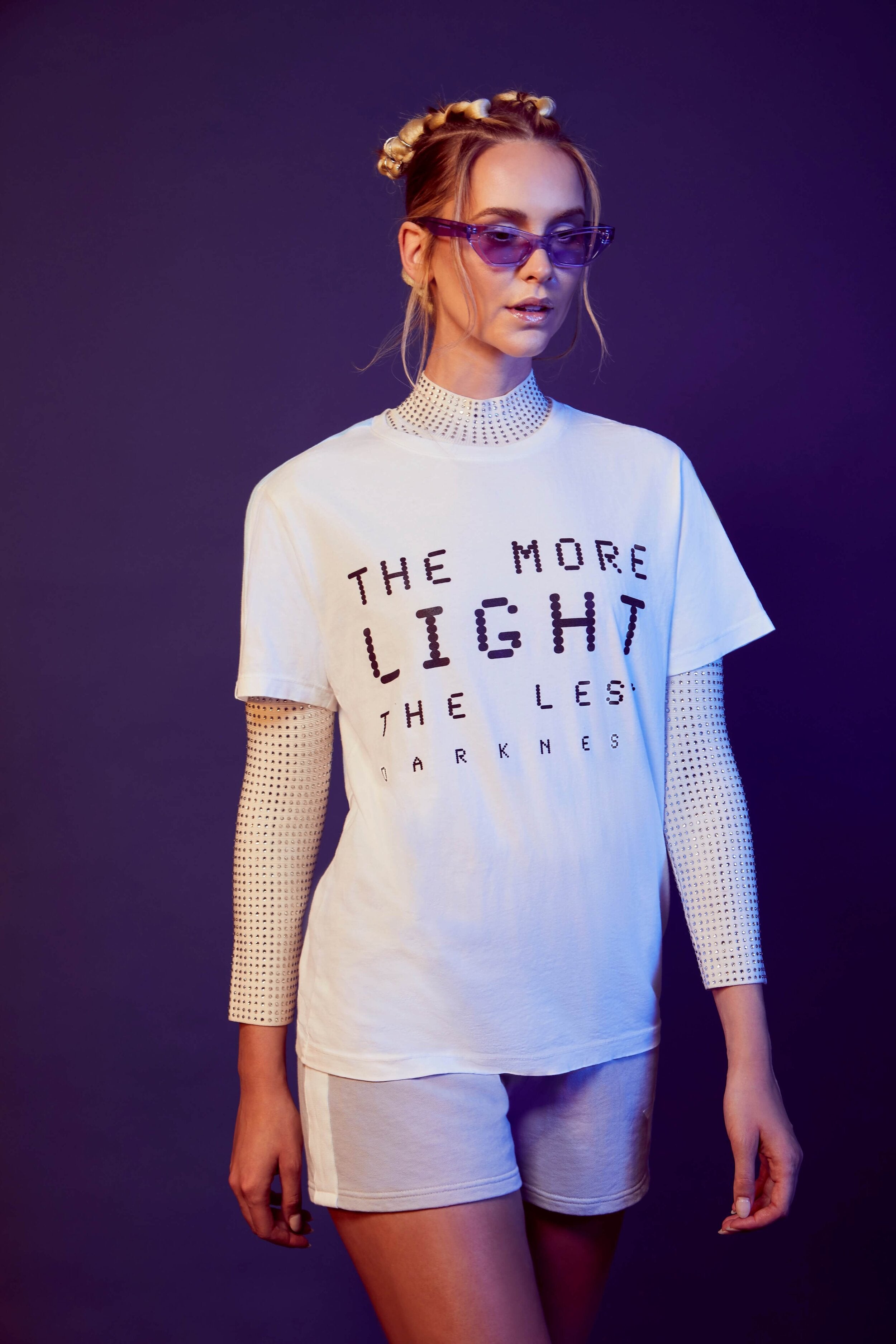 A person with blonde hair and sunglasses wears the GFLApparel More Light Tee in Lite Beam, layered over a white long-sleeve top and white shorts, standing against a dark background.