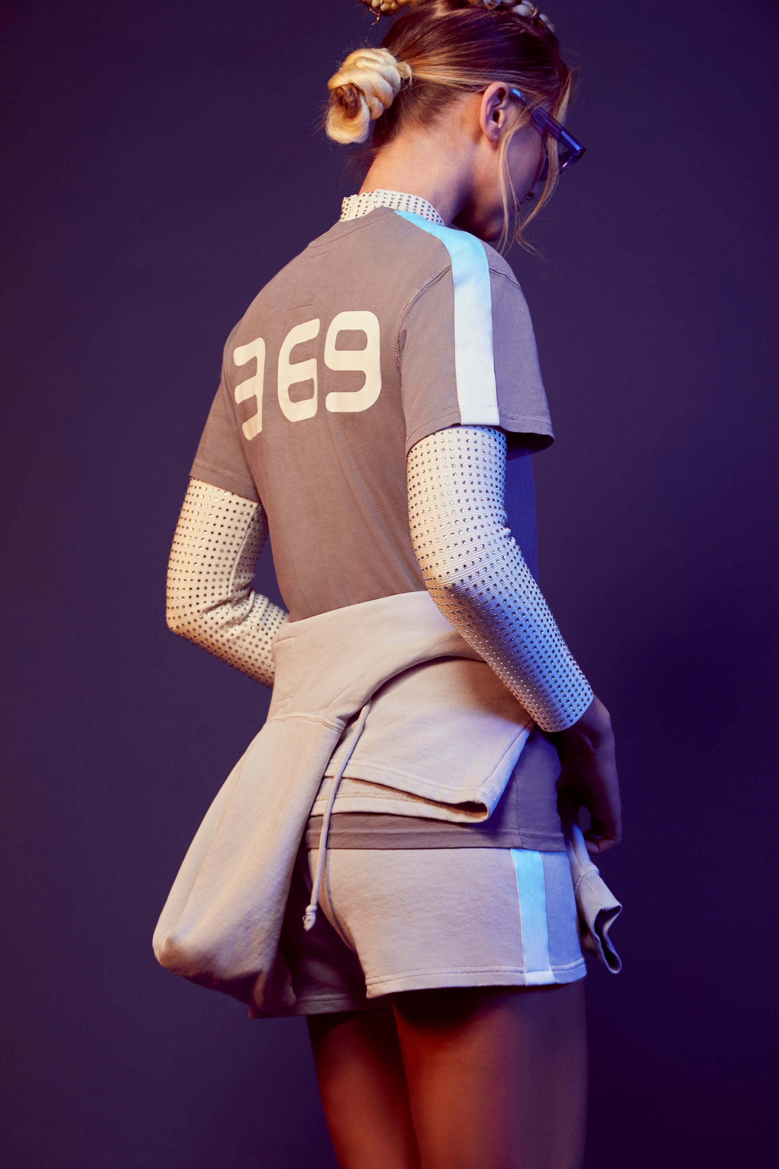 A person with blonde hair in a bun is standing and facing backward. They are wearing a grey 369 Tee in Asteroid from GFLApparel, reminiscent of Nikola Tesla's fascination with the number, over a white cotton jersey knit long-sleeve shirt, and have a grey sweatshirt tied around their waist.