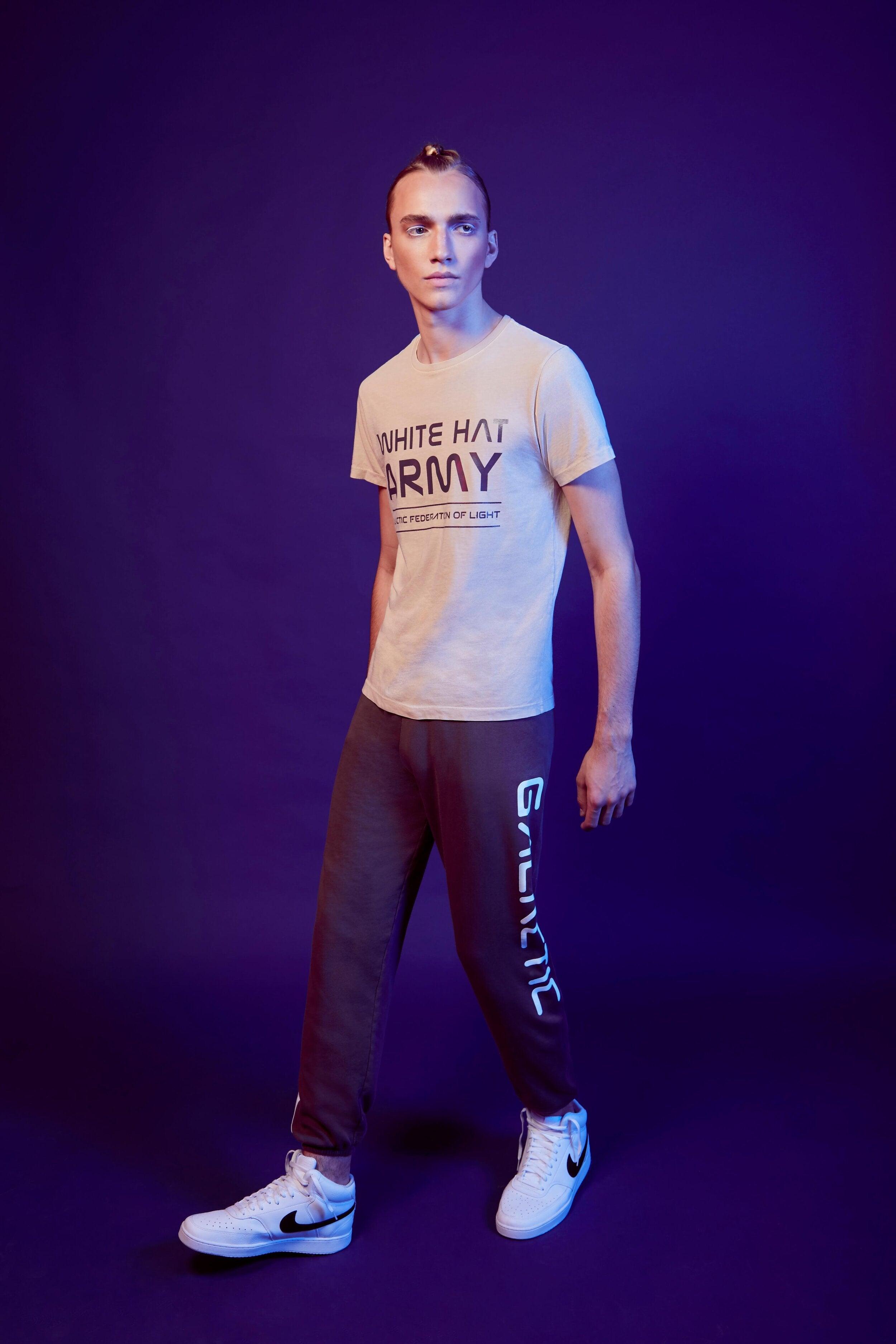 A person stands against a dark purple background, wearing a White Hat Army Tee in Dune by GFLApparel and dark sweatpants with text down the side. They are gazing into the distance and wearing white sneakers, making a bold statement with their look.