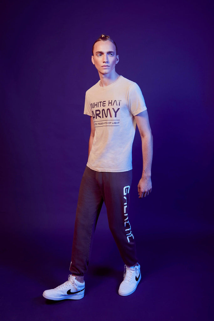 A person stands against a dark purple background, wearing a White Hat Army Tee in Dune by GFLApparel and dark sweatpants with text down the side. They are gazing into the distance and wearing white sneakers, making a bold statement with their look.