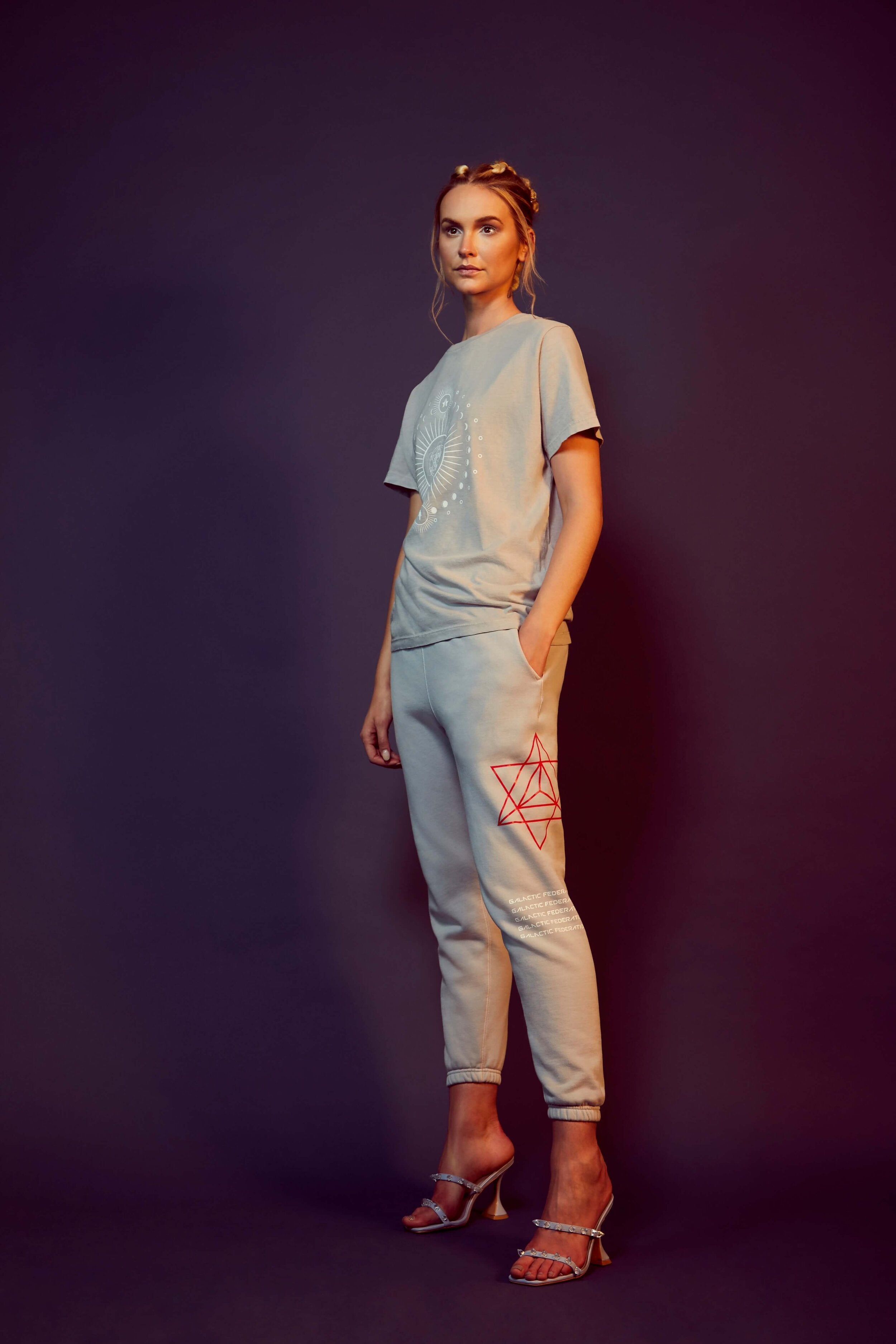 A woman stands against a dark background, wearing the GFLApparel Moon Metatron Tee in Galactic Gray and joggers adorned with red sacred geometric designs, and heels. She has her hair up and one hand in her pocket.