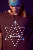 A person with long hair looks down, wearing a dark shirt made from breathable cotton. The shirt features a geometric design with overlapping white lines forming a layered star pattern, reminiscent of sacred geometry and the Merkaba. This is the Merkaba Tee in Space Glow by GFLApparel.