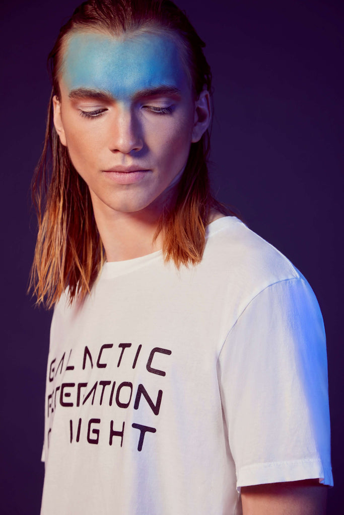 A person with long hair and a blue-painted forehead is wearing a GFL Stack Tee in Lite Beam by GFLApparel with the text "GALACTIC CITATION NIGHT" printed on it. They are standing against a dark background, embodying the spirit of the Galactic Federation of Light.