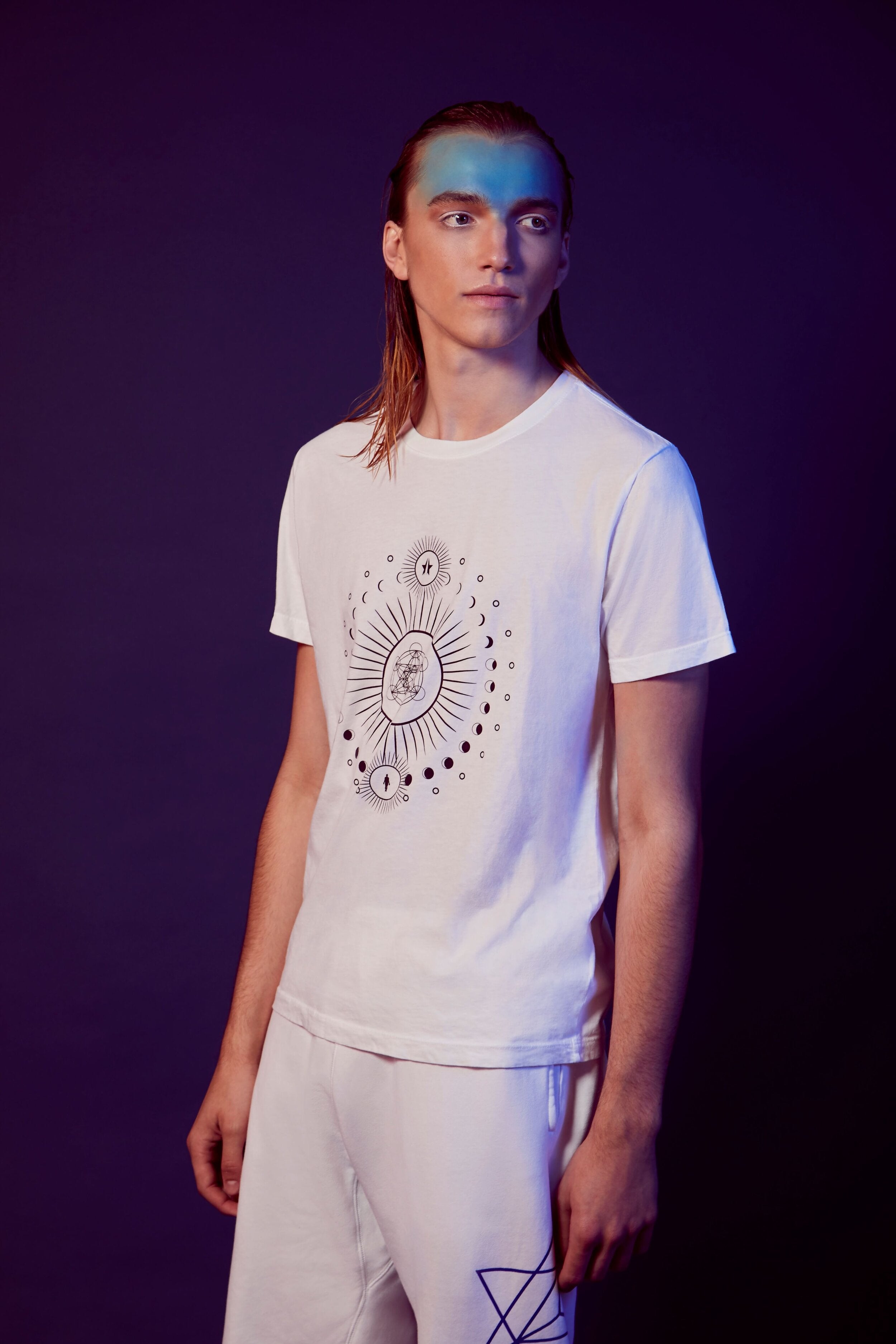 A person with long hair and blue face paint stands against a dark background, wearing the Moon Metatron Tee in Lite Beam by GFLApparel and white shorts.