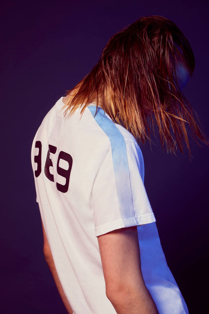 Person with long hair partially obscuring their face, wearing the GFLApparel 369 Tee in Lite Beam with the number '369' on the back, against a dark background—an homage to Nikola Tesla and his fascination with the sequence 3-6-9, often dubbed the key to the universe.