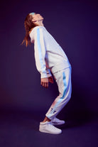 A person in a white tracksuit with blue stripes and a Merkaba Hoodie in Lite Beam from GFLApparel leans backward while balancing on one foot against a dark background.