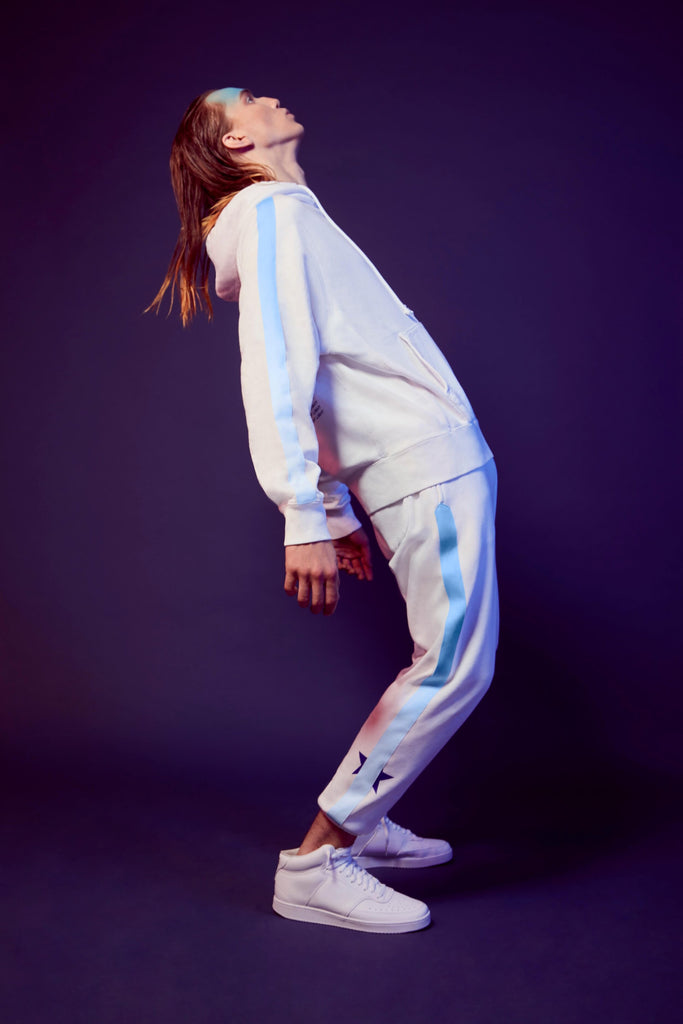 A person in a white tracksuit with blue stripes and a Merkaba Hoodie in Lite Beam from GFLApparel leans backward while balancing on one foot against a dark background.