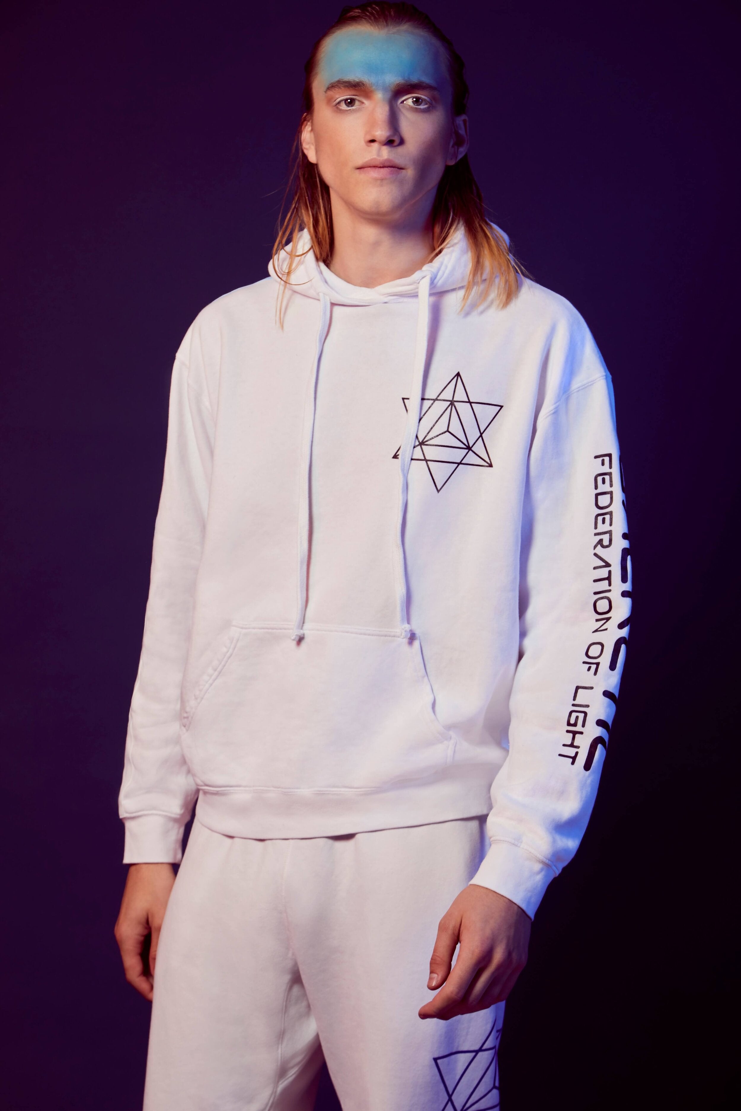 A person with long hair wears a Merkaba Hoodie in Lite Beam and matching sweatpants, both featuring sacred geometric designs and the text "Federation of Light" on the sleeve, against a purple background.