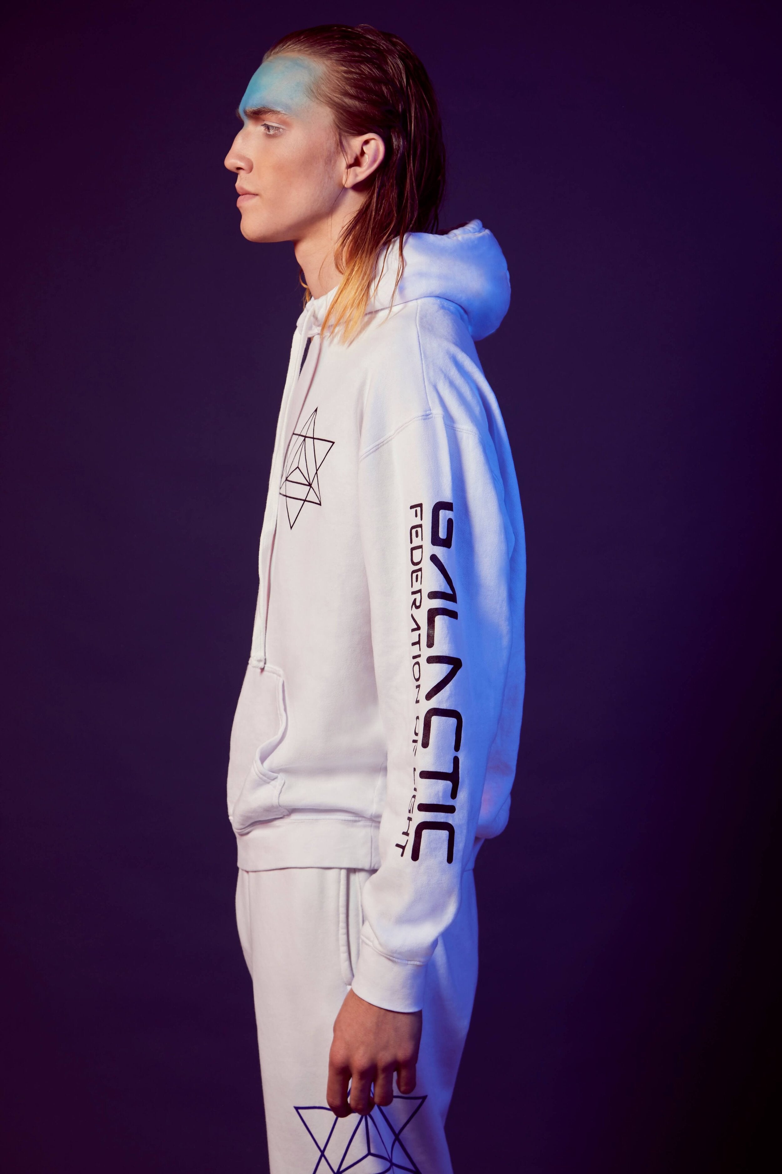 Person wearing a white **Merkaba Hoodie in Lite Beam** from **GFLApparel**, with "Galactic Federation" text on the sleeve and a Merkabah geometric design on the left chest and knee, standing in profile against a dark background.