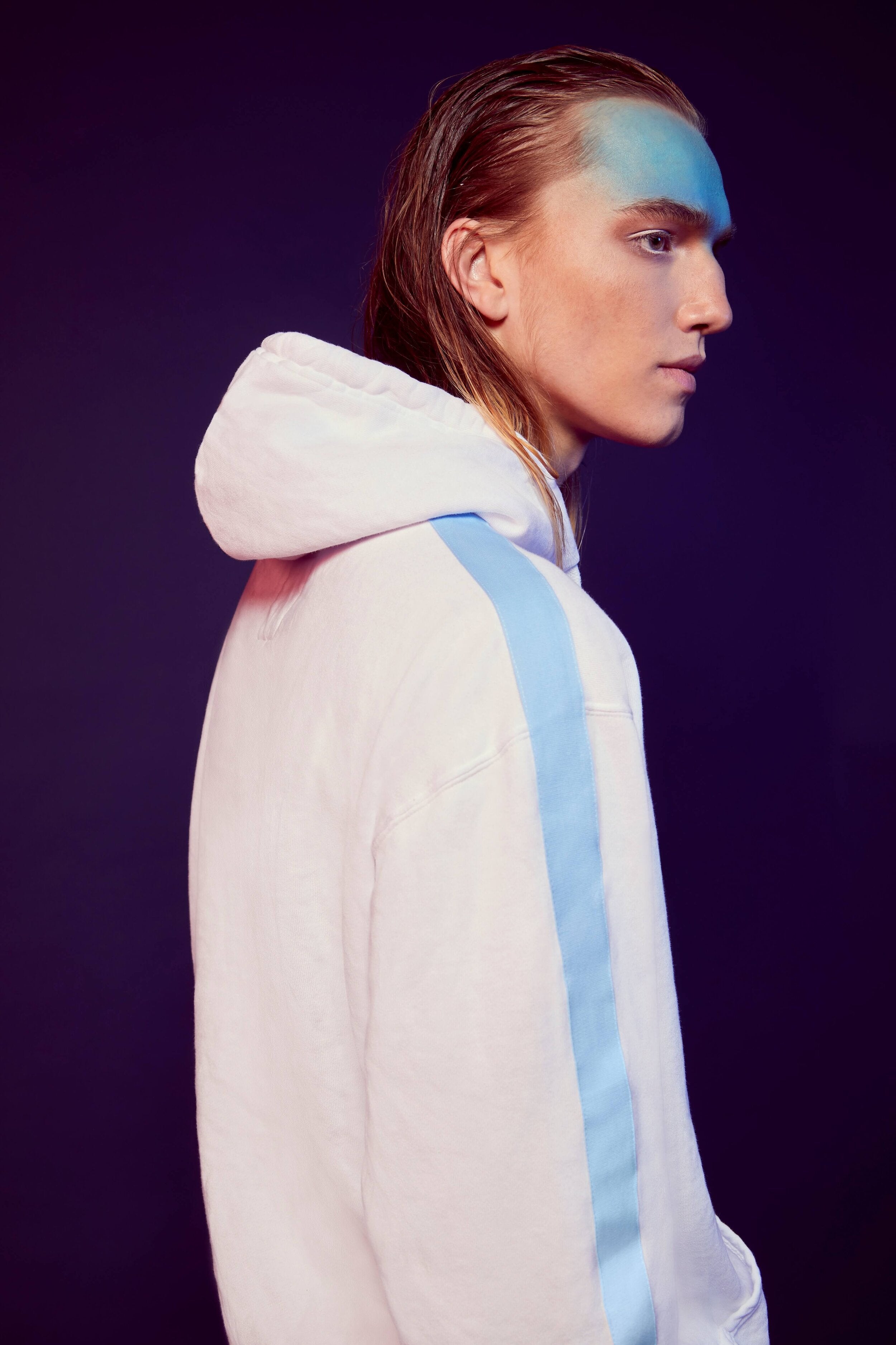 A person with shoulder-length hair is wearing a white GFLApparel GFL Stars Hoodie in Lite Beam with blue stripes, crafted from soft breathable cotton, and looking to the side against a dark purple background.