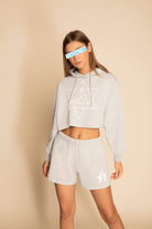 A person is wearing a light grey hoodie with bold print geometric designs and matching Star Shorts in Galactic Gray by GFLApparel. The background is neutral. The person's eyes are obscured with a pixelated pattern.