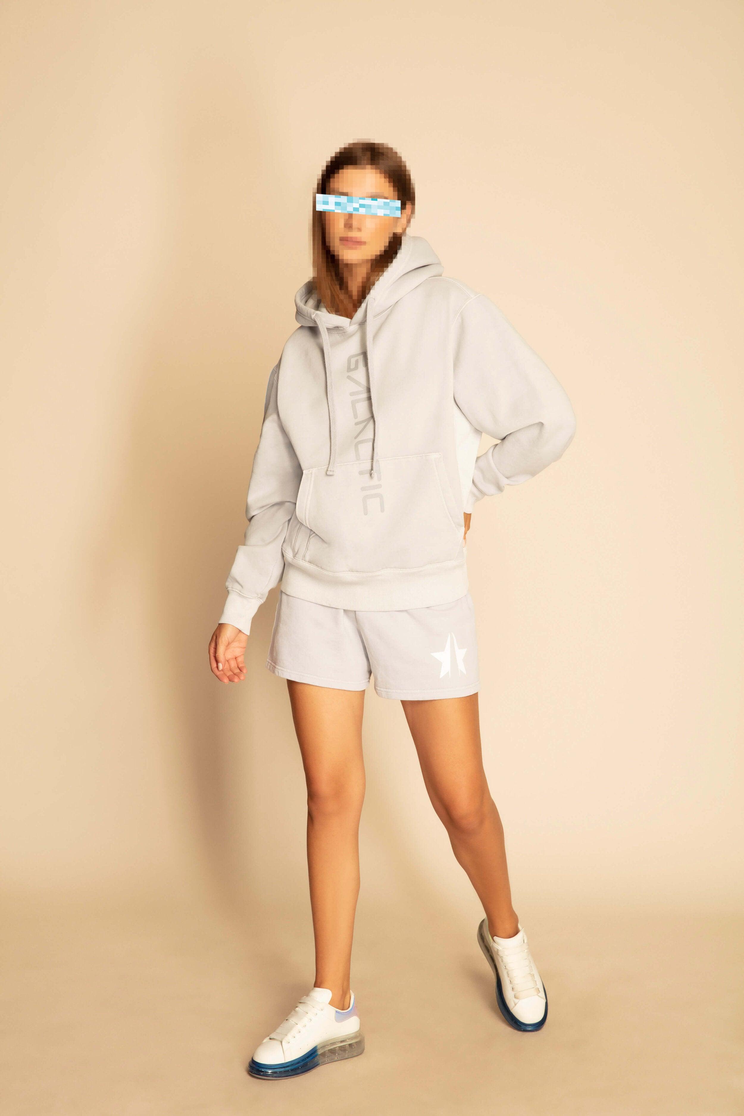 Person wearing a matching light gray French terry hoodie and GFLApparel Star Shorts in Galactic Gray, white sneakers, and sunglasses with a pixelated effect. They are standing against a plain background.
