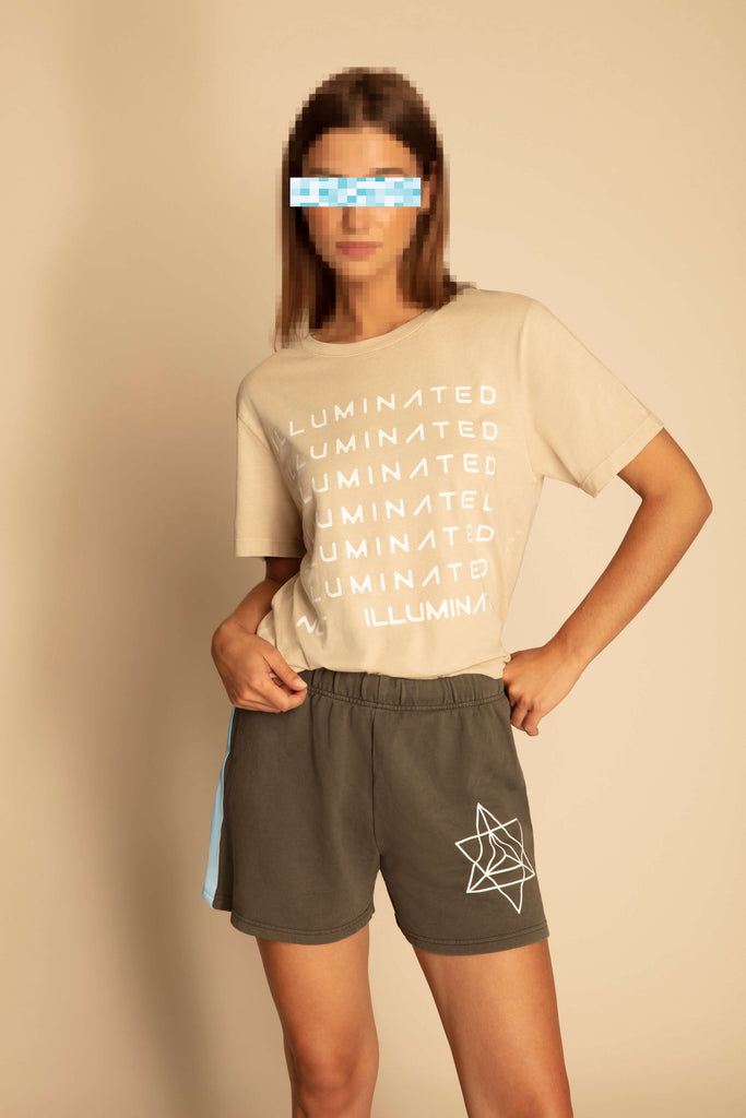 Person standing against a beige background wearing a soft and breathable cotton "Illuminated Tee in Dune" by GFLApparel in bold print, dark green shorts with a geometric design, and pixelation over their eyes.