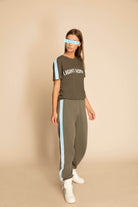 A person standing in a neutral pose wearing a dark green outfit with light blue stripes, white sneakers, and a breathable cotton GFLApparel Light Worker Tee in Caladan that reads "LIGHT WORK.