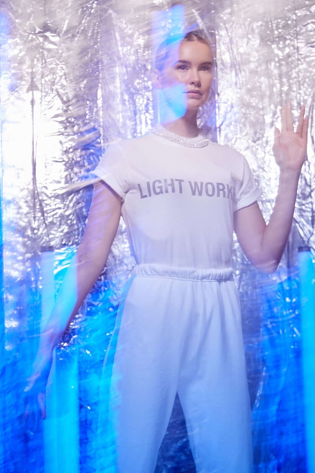 A person wearing a "Light Worker Tee in Lite Beam" by GFLApparel made of soft, breathable cotton and white pants stands against a reflective background with blue streaks of light.