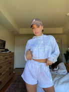 Person wearing a matching light-colored *Merkaba Cropped Hoodie in Galactic Gray* from *GFLApparel* and shorts set with a sacred geometric pattern on the hoodie, standing in a room with a dresser, TV, and bed in the background. A dog is lying on the bed.