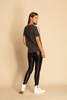 A person stands against a plain background, facing slightly sideways. They are wearing an Illuminated Tee in Space Glow from GFLApparel, black shiny leggings, and white high-top sneakers.