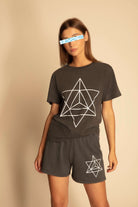 A person stands against a plain background, wearing a dark GFLApparel T-shirt and Merkaba Shorts in Space Glow with geometric star designs inspired by Sacred Geometry. Crafted from breathable cotton, their attire blends style and comfort seamlessly. For privacy, their face is pixelated.